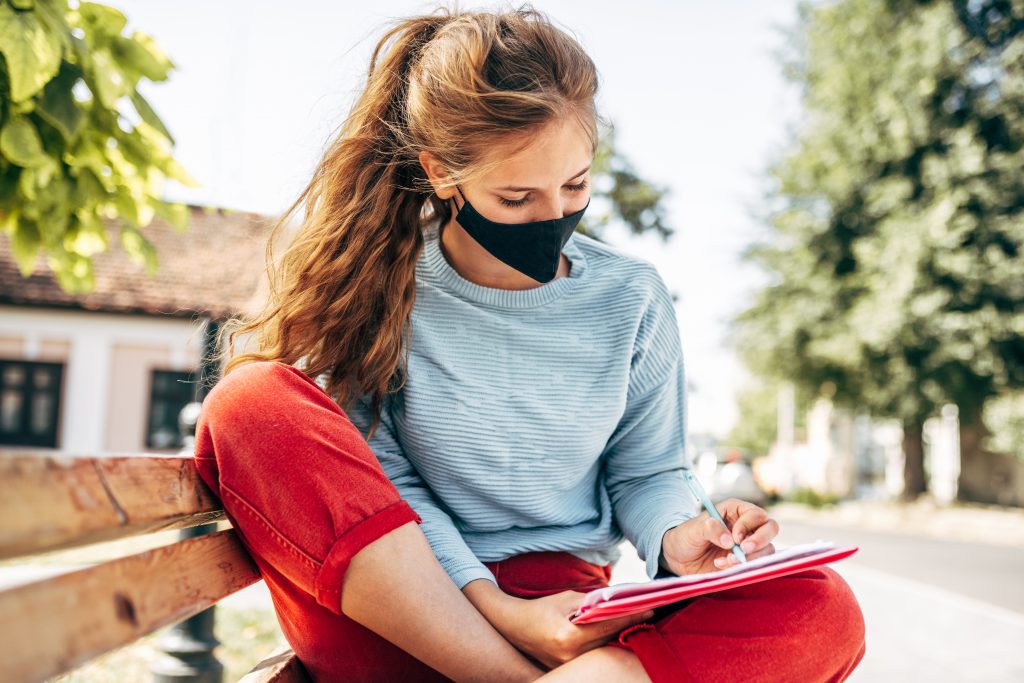 female student in a black protective face mask sitting on the bench learning outdoors on a sunny day.