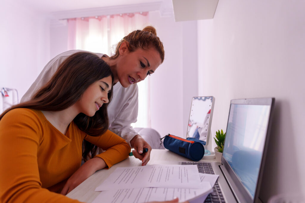 Mother and teenage daughter reviewing school papers and laptop at a desk, learning together for academic support and development