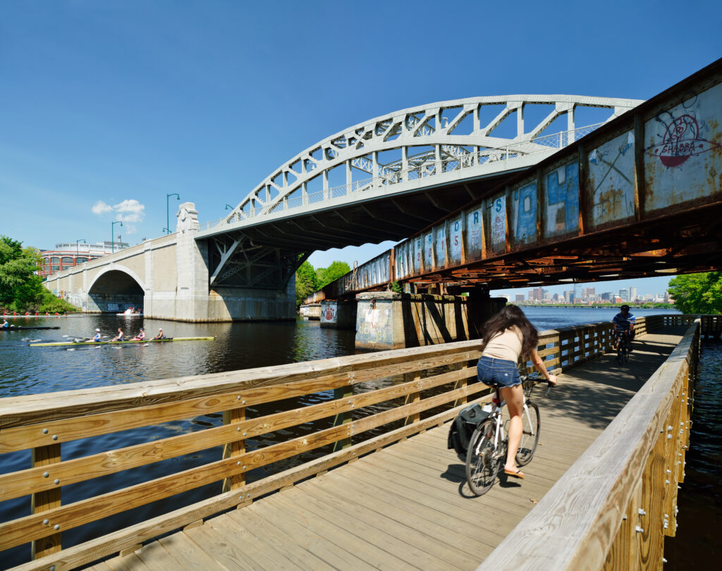 BU Bridge, old railroad and bike path on Charles River. People commuting and rowing, Boston city skyline in background.