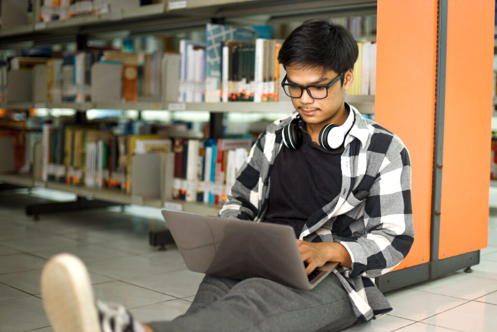 Young student leaning on library bookshelves while working on college essay on laptop