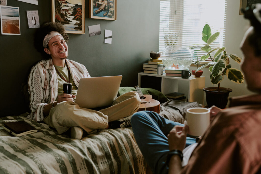 Curly-haired student sitting with laptop on bed and laughing out loud while his roomie telling joke and drinking coffee